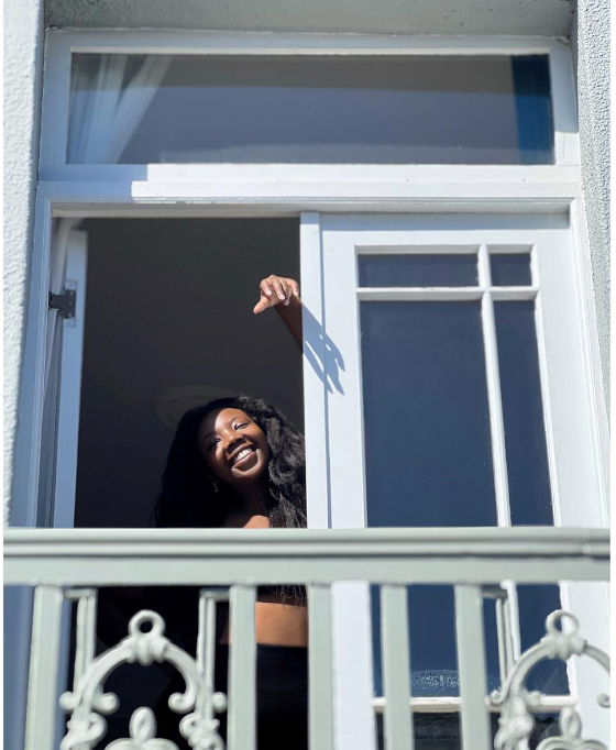 Ndiweni clicking photo standing by the balcony window smiling in the sun's embrace.